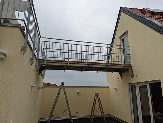Metallbrücke mit Geländer verbindet zwei beige Häuser über einem Innenhof mit zwei Leitern darunter