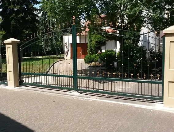 Grünes schmiedeeisernes Tor mit goldfarbenen Spitzen zwischen zwei hellen Pfeilern vor einem Garten mit Bäumen und einem Haus im Hintergrund