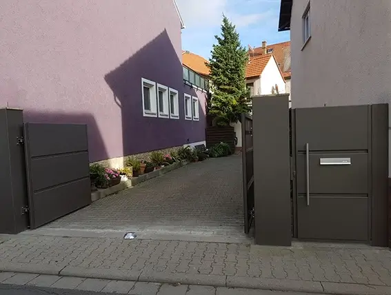 Offenes, modernes Einfahrtstor aus Metall vor gepflasterter Einfahrt zwischen zwei Häusern
